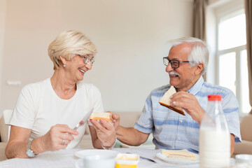 Fototapeta na wymiar Cheerful senior couple having breakfast at home. Elderly man hugging woman. Mature woman holding piece of bread. An old man and woman sitting at the table, talking.
