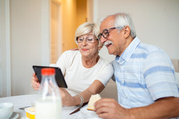 Senior man and his wife are using a digital tablet and communicating with their kids. Happy senior couple is having breakfast and having an online conversation with their family and sending greetings.