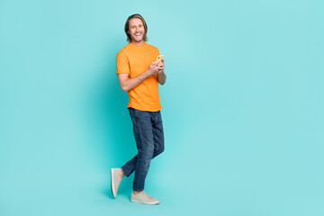 Full length photo of good mood guy dressed orange t-shirt chatting modern device empty space isolated teal color background