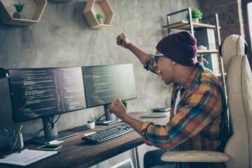 Photo of smiling lucky guy dressed eyewear hat rising fists successful start up indoors workstation...