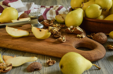 Closeup sliced pear pieces, split walnuts, kitchen mallet hammer on wooden cutting board indoors. Ripe yellowish pears in clay bowl on wooden table and traditional dishtowel on background