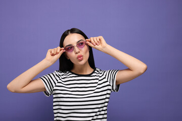 Beautiful young woman in stylish sunglasses blowing kiss on purple background