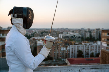 Portrait of young woman fencer wearing mask, white fencing costume and practicing outdoors.