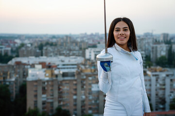 Portrait of young woman fencer wearing mask, white fencing costume and practicing outdoors.