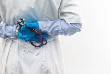 A doctor in a medical gown and blue gloves with a stethoscope in his hands in a hospital on a white background. Selective focus. Close-up