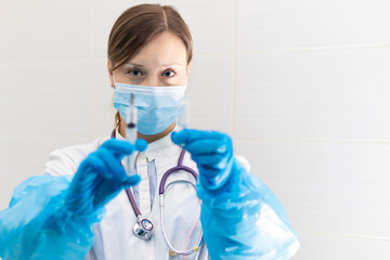 Young woman doctor in a medical mask and gloves with a syringe in her hand and a stethoscope in a hospital during a coronavirus pandemic. Selective focus. Portrait