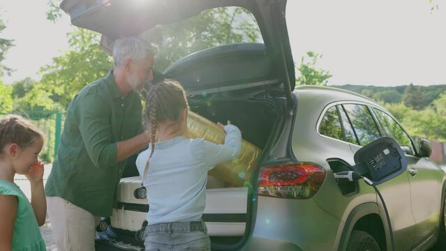 Father with kids packing suitcases into electric car trunk while charging battery.