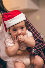 Mother is holding a baby in a Santa hat at home,close-up.Preparing for Christmas.Upcoming winter holidays and celebrations.