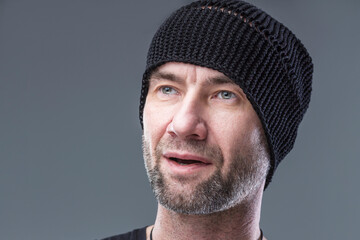 Brutal unshaven man in a black knitted hat. Grey background. Close-up.