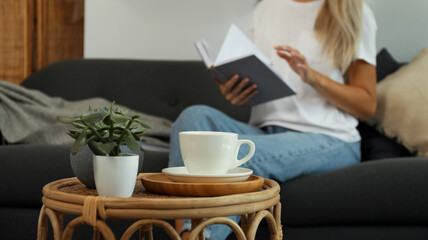 Woman reading book indoors, focus on table with cup and houseplants