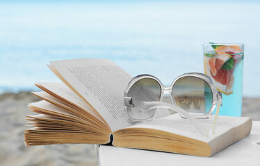 Open book, sunglasses and glass of refreshing drink on white table near sea