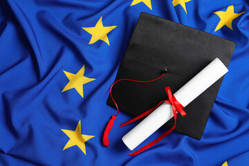 Black graduation cap and diploma on flag of European Union, flat lay. Space for text