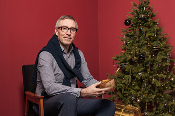 Attractive elderly man holds Christmas presents gold box against red studio wall background and green decorated Christmas tree