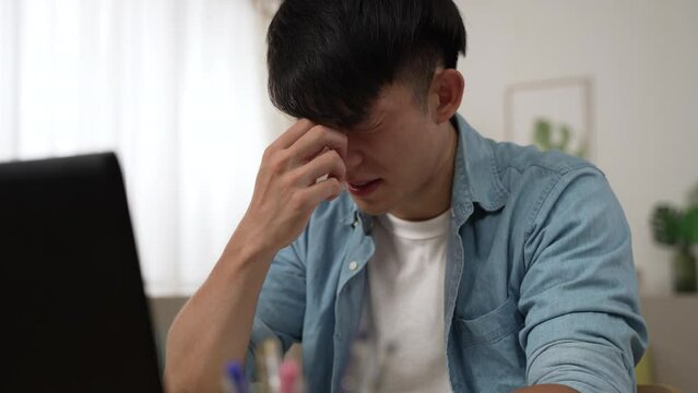 slow motion of a stressed asian male suffering a blurred vision is massaging his eyes and having trouble seeing clearly while working from home on the computer.