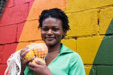 Smiling woman holding mesh bag with orange in front of colorful wall