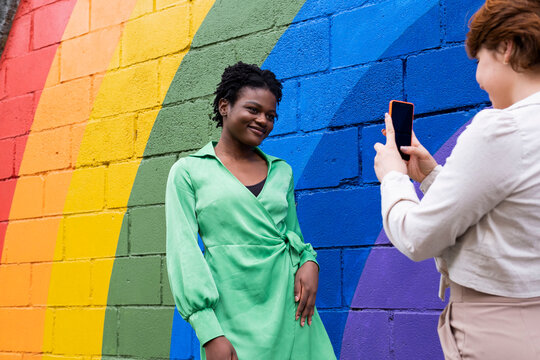 Smiling woman photographing girlfriend standing by rainbow flag painted on wall