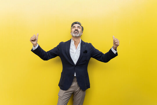 Cheerful businessman making thumbs up gesture standing against yellow wall