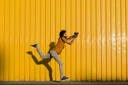 Man photographing through camera while running on footpath in front of yellow wall