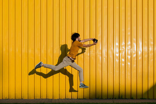 Man photographing through camera and jumping on footpath in front of yellow wall
