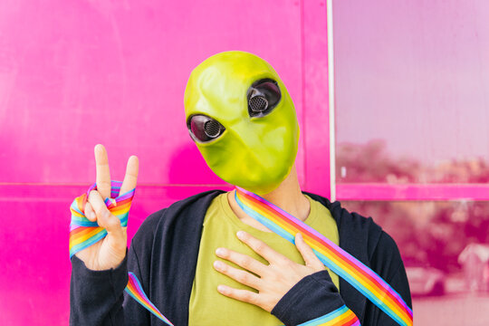 Man with alien mask and rainbow ribbon gesturing peace sign in front of pink wall