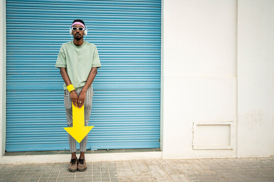 Man standing with arrow sign wearing headphones listening to music in front of closed blue shutter