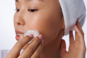 Closeup of asian woman with towel on head holding no-name jar of white cream. Young girl with a no-name jar of cream. Neutral natural pink manicure on nails. Woman doing cosmetic procedure