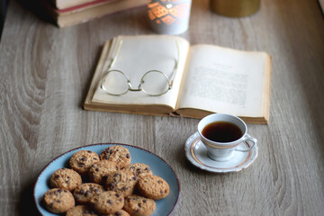 Plate of chocolate chip cookies, stack of vintage books, reading glasses, cup of tea or coffee, lit...