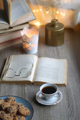 Plate of chocolate chip cookies, stack of vintage books, reading glasses, cup of tea or coffee, lit candle and fairy lights. Hygge at home. Selective focus.