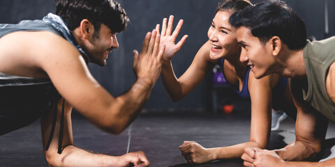 Young fit and health conscious group of male and female friends clapping hands while practicing single hand balance planks pose in modern dark gym floor in sportswear while training together