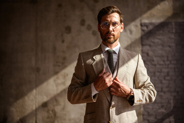 Stylish businessman in eyeglasses and suit looking at camera while standing in sunlight in office 