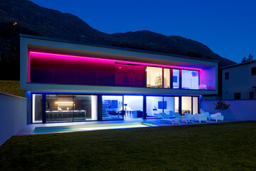 Modern house with swimming pool and garden in night scene illuminated by colored LED lights. Behind the house is the hill with the forest