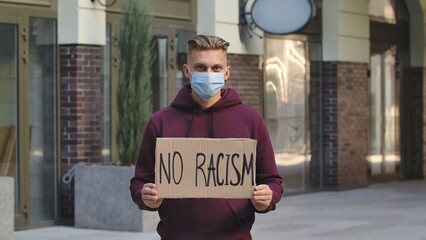 A young and blond man protester activist is wearing a medical mask holds a cardboard poster with...