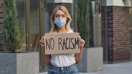 Caucasian female protester activist wearing a medical mask holds a cardboard poster with the slogan...