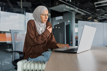 Businesswoman freezing in office at work, woman in hijab near heater trying to keep warm working...