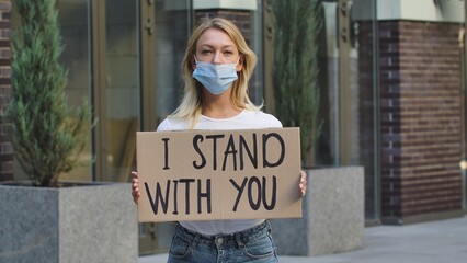 Caucasian female protester activist wearing a medical mask holds a cardboard poster with the slogan I STAND WITH YOU. A protest against racism and violence during quarantine. Stop racism concept.