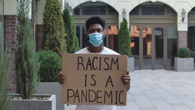 Young African American Man In A Medical Mask Stands With A Cardboard Poster RACISM IS A PANDEMIC In A Public Outdoor Place. An Anti Racist Movement To Protest Against Injustice. Street Demonstration.