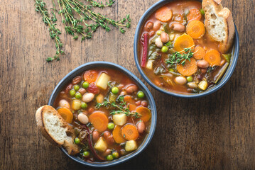 Minestrone vegetable soup on a wooden textured background with pieces of toasted bread and herbs. Simple vegan rustic Italian dish, classic of Italian cuisine, delicious healthy Mediterranean lunch