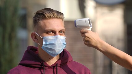 Portrait of young man in medical mask whose temperature is measured with contactless thermometer....