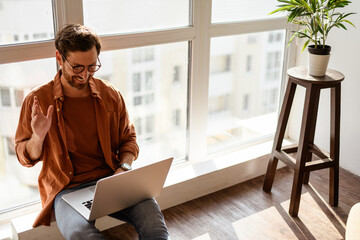 Smiling man in eyeglasses having video call on laptop on windowsill in studio 