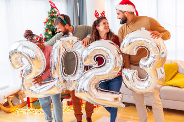 Friends holding big balloons shaped as numbers 2023 while celebrating New Year at home