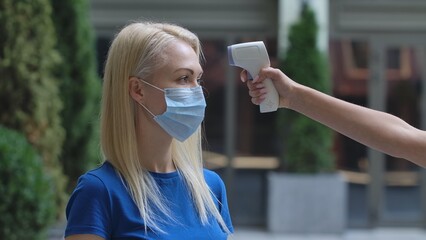 Portrait of young woman in medical mask whose temperature is measured with contactless thermometer....