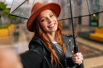 Smiling redhead young woman wearing elegant hat under transparent umbrella making selfie photo looking at camera, holding mobile phone on hand, standing in rain on European city street