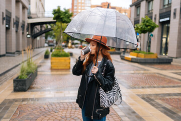 Happy attractive young woman wearing fashion hat standing with transparent umbrella in city cold...