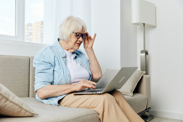 an elderly woman is carefully peering at the laptop monitor trying to read the information holding her glasses with her hand holding it on her lap while sitting on the couch