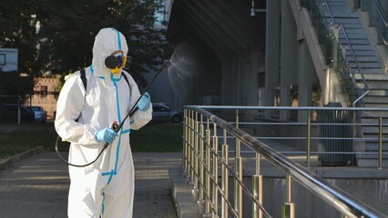 Medical working man in a white protective suit and respirator disinfects, sprays chemicals on surface railing. Treating objects with an antiseptic using an alcohol disinfectant and sprinkler.