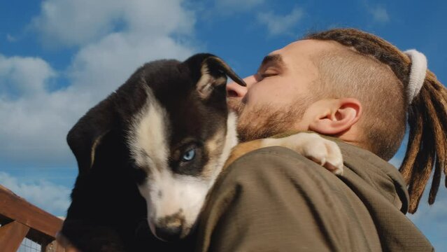 Young handsome Caucasian man with dreadlocks came to dog shelter to choose new friend. Volunteer plays blue eyed Alaskan husky puppy. Concept adopting pets. Close up portrait against blue cloudy sky.