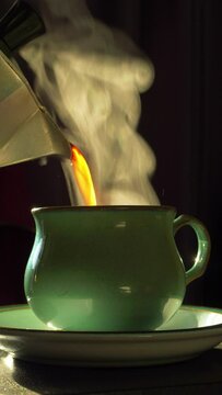 Vertical video social media format – Closeup isolated shot of backlit steaming hot coffee being poured from a traditional stovetop moka pot into a cup on a saucer.