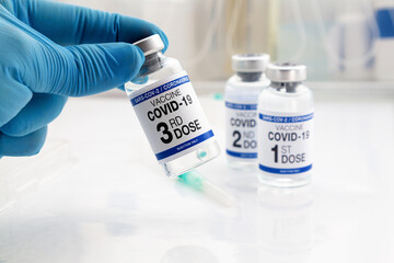 Hand holding COVID-19 Vaccine Vial for vaccination tagged with 3rd dose and background 1st, 2nd...