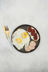 Balanced breakfast of two fried eggs, sandwich of cream cheese and dried tomatoes, slices of poultry meat on gray background.
