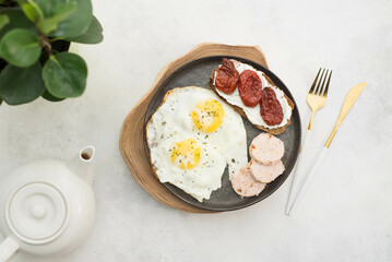 breakfast of two fried eggs, sandwich of cream cheese and dried tomatoes, slices of poultry meat with tea pot on gray background.